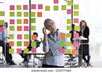 Thoughtful Middle Aged Office Employee Woman Thinking Over Kanban Board. Senior Business Lady Analyzing Project Tasks, Strategy, Plan, Looking At Paper Notes On Glass. Scrum Management Concept