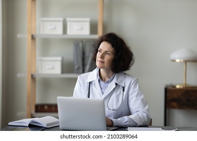 Thoughtful Middle Aged Old Female General Practitioner Doctor Sitting At Table With Laptop, Looking In Distance Considering Healthcare Problem Solution Or Difficult Illness, Working In Clinic Office.