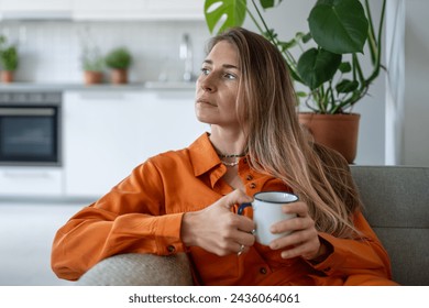 Thoughtful middle aged woman sits on sofa at home holding cup in hand looks towards window daydreaming. Peaceful pensive female thinking about future in cozy apartment in moment of quiet introspection - Powered by Shutterstock