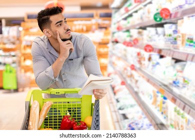 Thoughtful Middle Eastern Man Doing Grocery Shopping Calculating Expensive Prices Holding Shopper List And Looking Aside At Shelf With Food Products Standing With Shop Cart In Supermarket