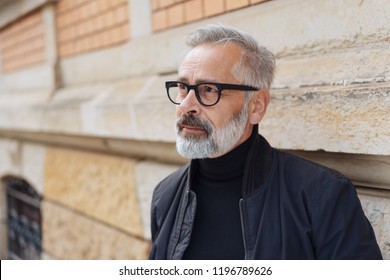 Thoughtful middle-aged bearded man wearing glasses standing outdoors in town against a receding wall with copy space - Powered by Shutterstock