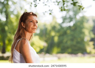 A thoughtful, middle-aged woman wearing a white dress contemplates quietly in a lush, green park bathed in sunlight. - Powered by Shutterstock