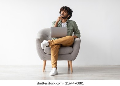 Thoughtful Millennial Eastern Man Sitting In Armchair, Using Brand New Computer, Unemployed Indian Guy Seeking Job On Internet, Looking At Copy Space, White Studio Background