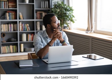 Thoughtful mixed race businessman sitting at table with computer, looking away. Distracted from study job young african american man lost in thoughts, thinking of difficult decision at home office.