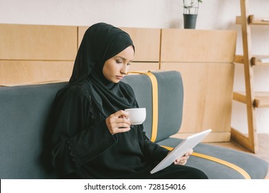 thoughtful muslim woman drinking coffee and looking at digital tablet