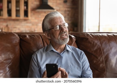 Thoughtful old age man sit on couch hold phone look aside distracted from screen recall in memory contact of friend to make call. Worried hoary senior grandpa in glasses need help in using mobile app