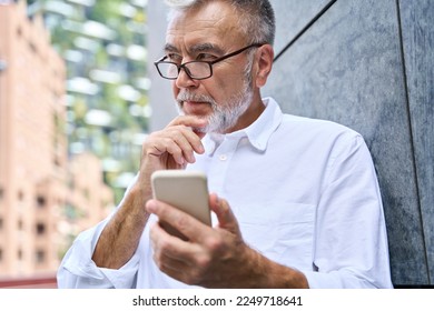 Thoughtful old bearded mature adult professional business man, senior older businessman wearing glasses holding mobile using cell phone thinking of business ideas standing outdoors. - Powered by Shutterstock