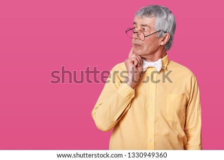 Thoughtful old man with black rounded glasses on nose. Elderly senior looks aside and keeps right hand on chin. Grey haired pensioner thinks about something interesting isolated over rose background.