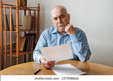 Thoughtful Old Man Reading A Handheld Paper Document With A Look Of Concentration As He Sits Indoors At A Table