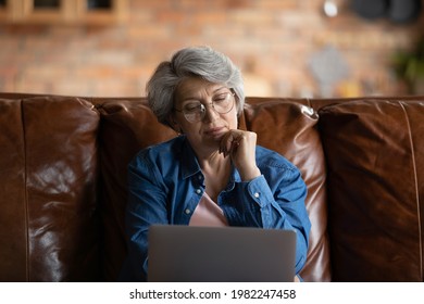 Thoughtful Old Woman In Glasses Sit On Sofa With Laptop, Touch Chin With Hand Having Doubts Looks Puzzled, Feels Confused Need Help For App Understanding, Experiences Troubles With Modern Tech Concept