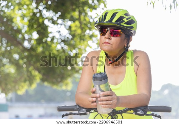 Thoughtful old woman in protective\
sportswear leaning on bicycle handlebar with water\
bottle