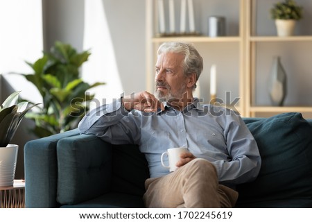 Thoughtful older man sitting on couch at home alone, holding cup of tea or coffee, feeling lonely and sad, unhappy depressed mature male looking into distance, thinking about problems