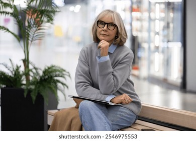 Thoughtful older woman sitting in a modern shopping mall, surrounded by greenery, reflecting on her purchases during a quiet afternoon - Powered by Shutterstock