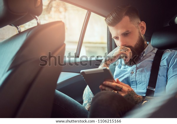 Thoughtful old-fashioned tattooed hipster guy in a
shirt with suspenders, using a tablet while sitting in a luxury car
on back seat.