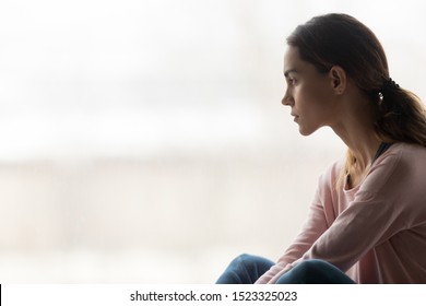 Thoughtful pensive girl sitting alone looking through window thinking of problem or reflecting, anxious melancholic young woman serious face lost in thoughts feel lonely doubtful worried, side view