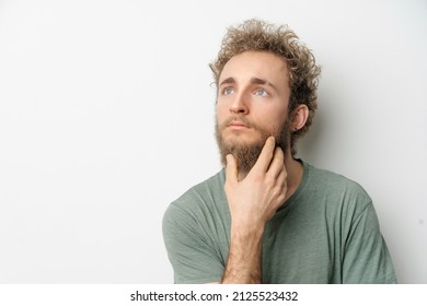 Thoughtful or pensive young handsome bearded wild curly hair man with bright blue eyes isolated on white background. Young thinking man in green t shirt on white. Copy space.  - Powered by Shutterstock