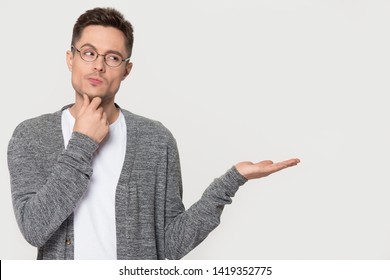 Thoughtful Pensive Young Man In Glasses Touch Chin Thinking Standing On Grey White Background Isolated Studio Shot Showing Empty Copy Space On Open Hand Palm Feels Unsure Doubtful Concept Image