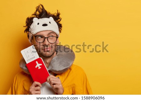 Thoughtful plane passenger prepares documents and flying tickets on boarding pass, thinks about next travel, has tired look, slept in transport, looks aside, wears blindfold, glasses, neck pillow