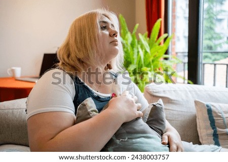 Thoughtful plus-size woman sits on ouch at home, clutching a cushion, with somber expression amidst indoor plants.