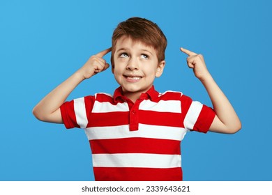 Thoughtful preschool boy in red striped polo shirt pointing at head and looking up while thinking against blue background. - Powered by Shutterstock