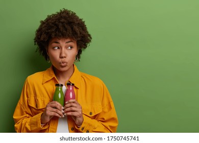 Thoughtful Pretty Afro American Woman Holds Glass Bottles Of Fresh Apple And Strawberry Smoothie, Leads Healthy Lifestyle, Drinks Nutrient Organic Beverage To Keep Fit, Isolated On Green Background