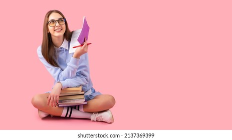 Thoughtful pretty blond school girl looking up with a smile sitting on the floor with crossed legs holding books and a diary with a pen, high school university concept. - Shutterstock ID 2137369399