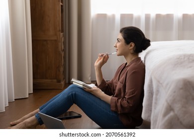 Thoughtful pretty young Indian woman sit in bedroom on floor holds pen thinks over future business plan, take notes in paper notebook, write to-do list creative ideas looking inspired, study concept
