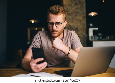 Thoughtful Redhead Man Of 30 Years Old Using Smartphone App While Sitting Front Open Laptop Computer Indoors. Puzzled Male Reading Message On Mobile Phone During Freelance Work In Coworking Space