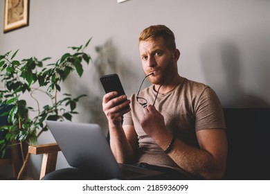 Thoughtful Redhead Man Of 30 Years Old Using Smartphone App While Sitting Front Open Laptop Computer Indoors. Puzzled Male Reading Message On Mobile Phone During Freelance Work In Coworking Space