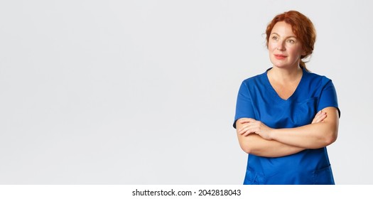 Thoughtful Redhead Nurse, Physician Or Female Doctor In Scrubs Looking Upper Left Corner With Intrigued, Interested Expression, Cross Arms Chest, Turn Attention To Banner, Grey Background