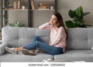 Thoughtful relaxed woman in casual clothes rest on comfy sofa in cozy modern living room looks out the window enjoying view or daydreaming, girl smiling lost on pleasant thoughts spend weekend at home