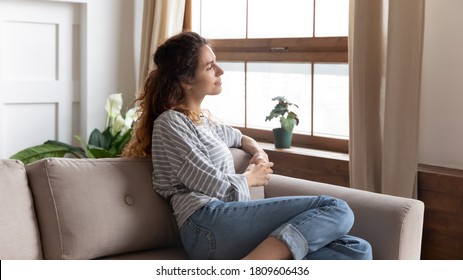 Thoughtful relaxed woman in casual clothes rest on couch in cozy modern living room looks out the window enjoy city admires view, daydreaming, smiling lost on pleasant thoughts spend weekend at home