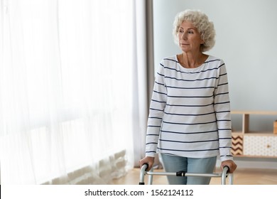 Thoughtful Sad Disabled Injured Old Woman Standing Alone Holding Walking Frame At Home, Depressed Lonely Handicapped Senior Grandmother Look Through Window Using Walker, Elder People Physiotherapy