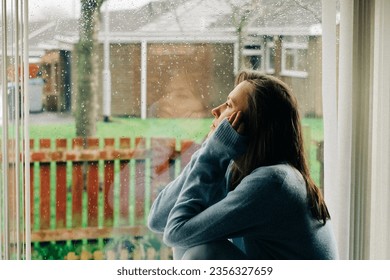 Thoughtful sad woman sitting on a windowsill with a reflection on a window. Mental health and seasonal disorder due to rainy cold weather, end of summer concept