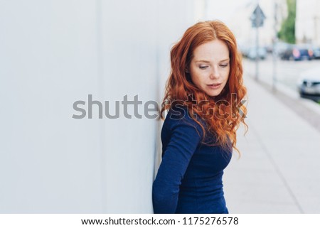 Thoughtful sad young woman with downcast eyes leaning against a white exterior wall in a town street with copy space