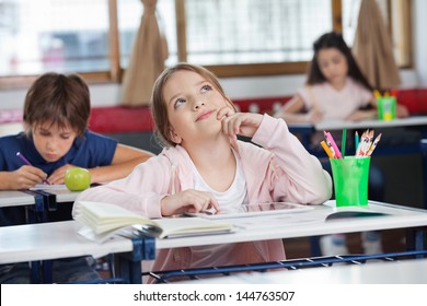 Thoughtful schoolgirl with digital tablet looking up while friends studying in background at classroom - Powered by Shutterstock