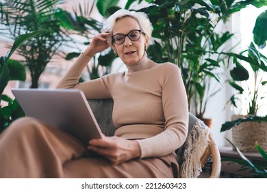 Thoughtful Self Employed Woman With Short Gray Hair Sitting On Chair And Using Laptop While Working Remotely On Business Startup In Living Room At Home