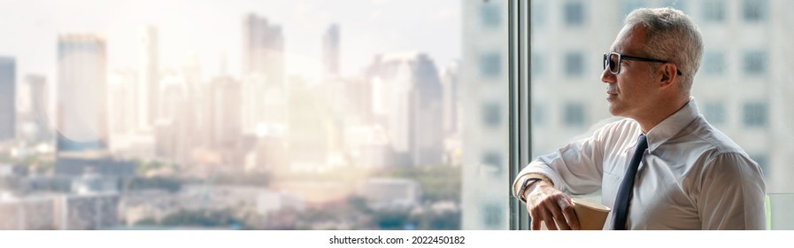 Thoughtful Senior Business Man Holding Cup Of Coffee Standing Over Window Glass In Office On High Building Cityscape View Background . Worry Old Worker Distracted . Executive Thinking And Consider 