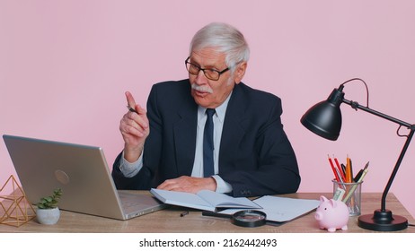 Thoughtful Senior Businessman Making Notes, Writing Down Thoughts With Pen Into Notepad Notebook Diary, To Do List, Good Idea Sits At Workplace Desk At Office. Elderly Man On Pink Studio Background