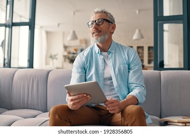 Thoughtful senior man using digital tablet and looking away with smile while sitting on the sofa at home - Powered by Shutterstock