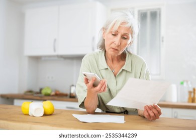 Thoughtful senior woman analyzing bills and using smartphone, standing at kitchen table at home. - Powered by Shutterstock