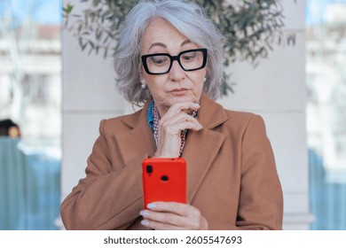 Thoughtful senior woman with gray hair and glasses using a smartphone outdoors. Digital communication, modern lifestyle, and technology concept in a stylish urban setting with natural light. 