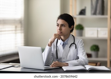 Thoughtful serious Indian female doctor physician touching chin sitting at work desk in office with laptop, looking in distance, pensive practitioner in white uniform with stethoscope thinking