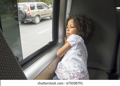 Thoughtful And Serious Little Girl Looking Out The Window In The Bus. Small Tired And Sad Female Mixed Ethnicity Child With Curly Hair Sitting On The Bus Chair