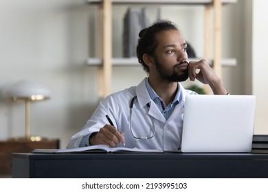 Thoughtful serious male doctor looking at window in office, thinking of patients, future vision, medical career growth, promotion, working at table with laptop, writing records.