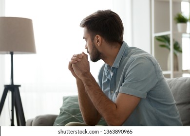 Thoughtful serious man sitting on sofa alone at home, lost in thoughts, thinking about problem solving, feeling lonely, making important decision, having psychological problem, side view