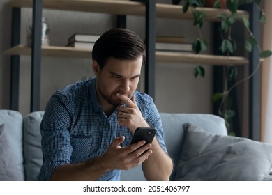 Thoughtful serious man touching chin, looking at smartphone screen, sitting on couch at home, pensive young businessman or student reading bad news in message, unexpected debt, solving problem