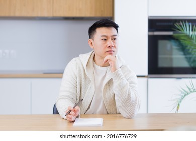 Thoughtful Serious Young Asian Man Looking Away. Student Writer Sit At Home Office Desk With Laptop Thinking Of Inspiration Search Solution Ideas Lost In Thoughts Dreaming Solving Financial Problems