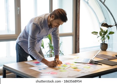 Thoughtful serious young businessman employee standing at desk focused on paperwork, making changes correcting mistakes in marketing plan by hand, analyzing client customer demands using infographics