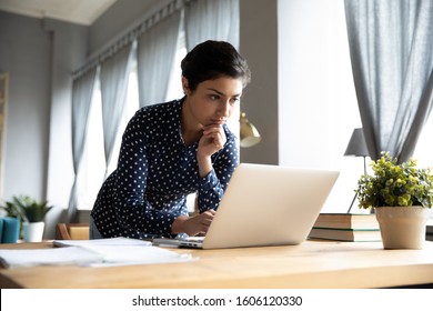 Thoughtful serious young indian ethnic woman student freelancer working studying on laptop computer looking at pc screen focused on thinking solving online problem doing research at home office desk.